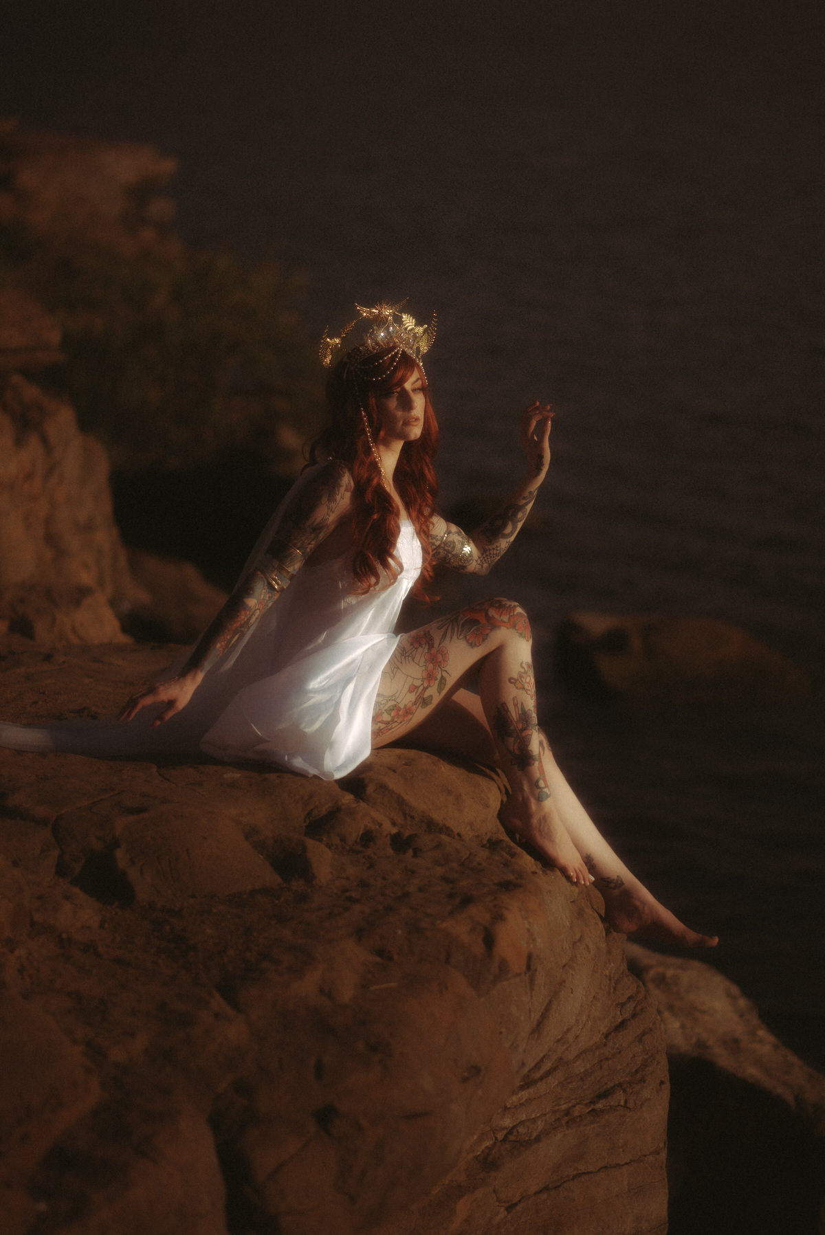 Woman with red hair in a white slip dress and wearing a crown sitting on a cliff at the ocean.