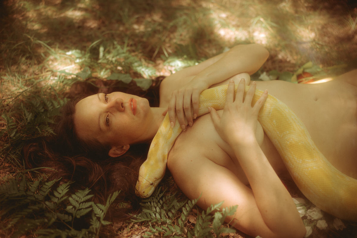 A naked woman laying down in the forest with vines and ferns surrounding her while holding a large snake around her body.