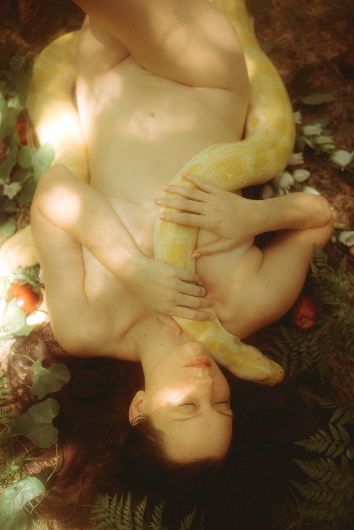 A woman hugging a snake to her naked body while laying in the forest.