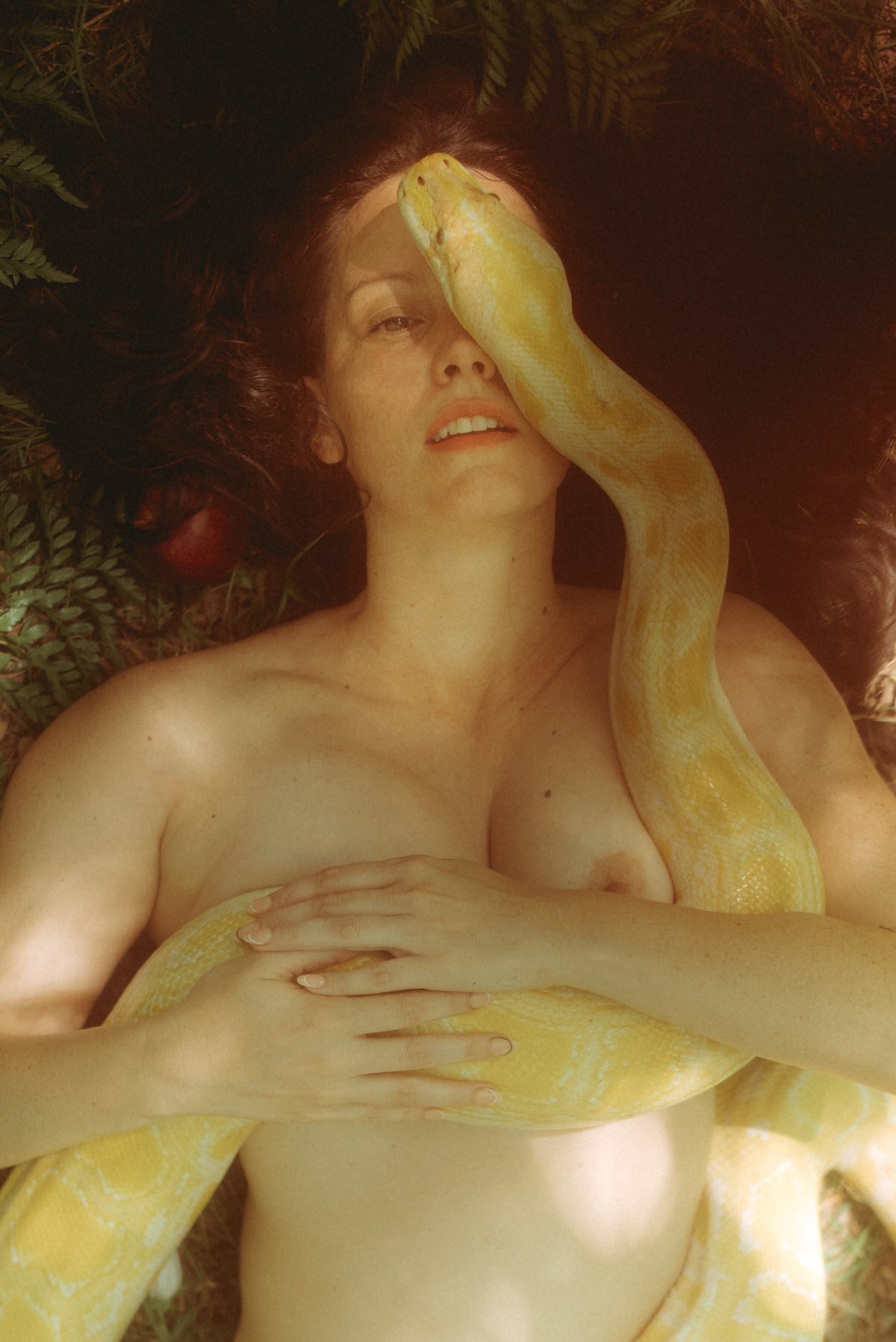 A woman laying on her back in the forest with a snake covering half of her face.