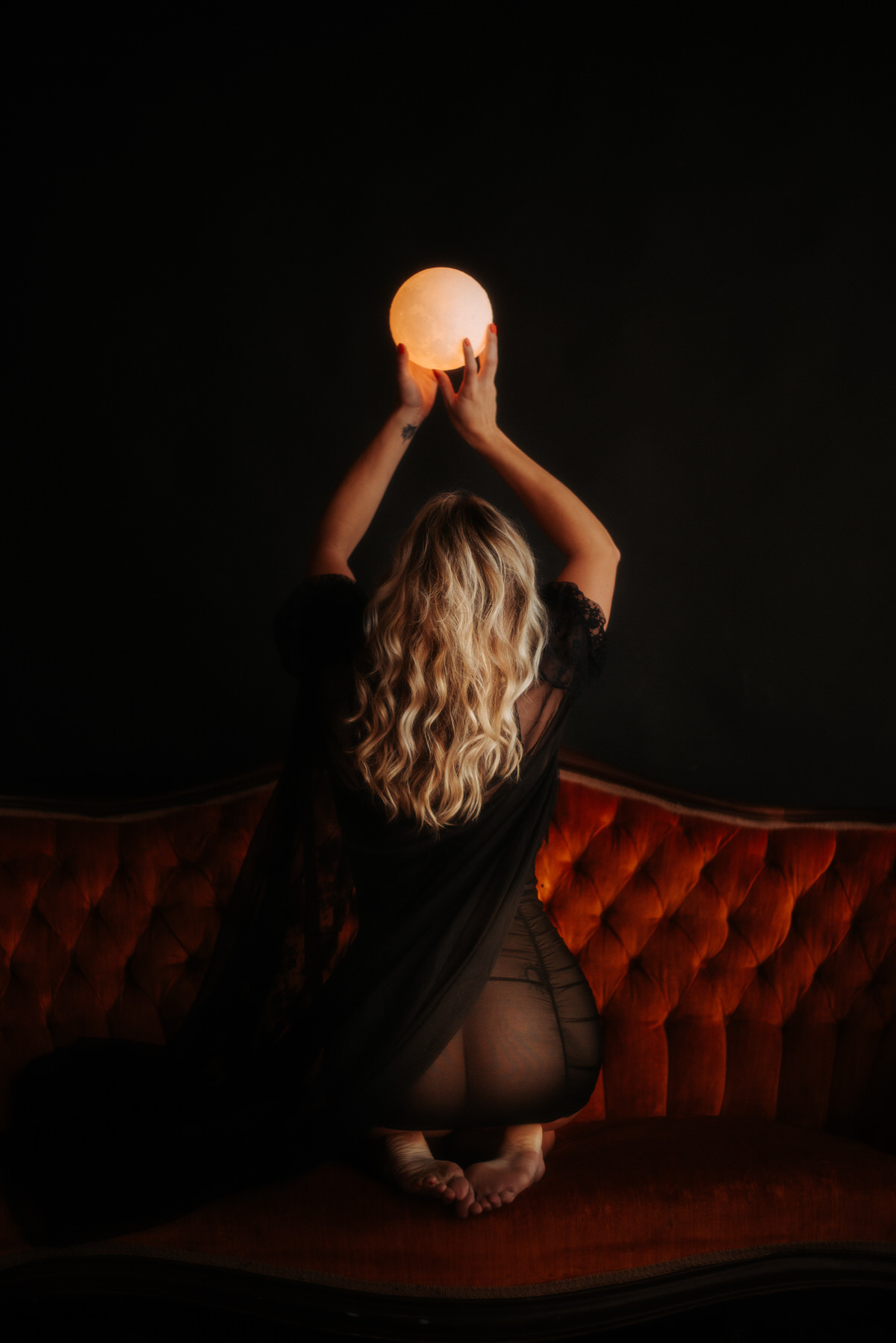A woman with blonde hair kneeling on an orange couch while holding up a moon lamp above her head.
