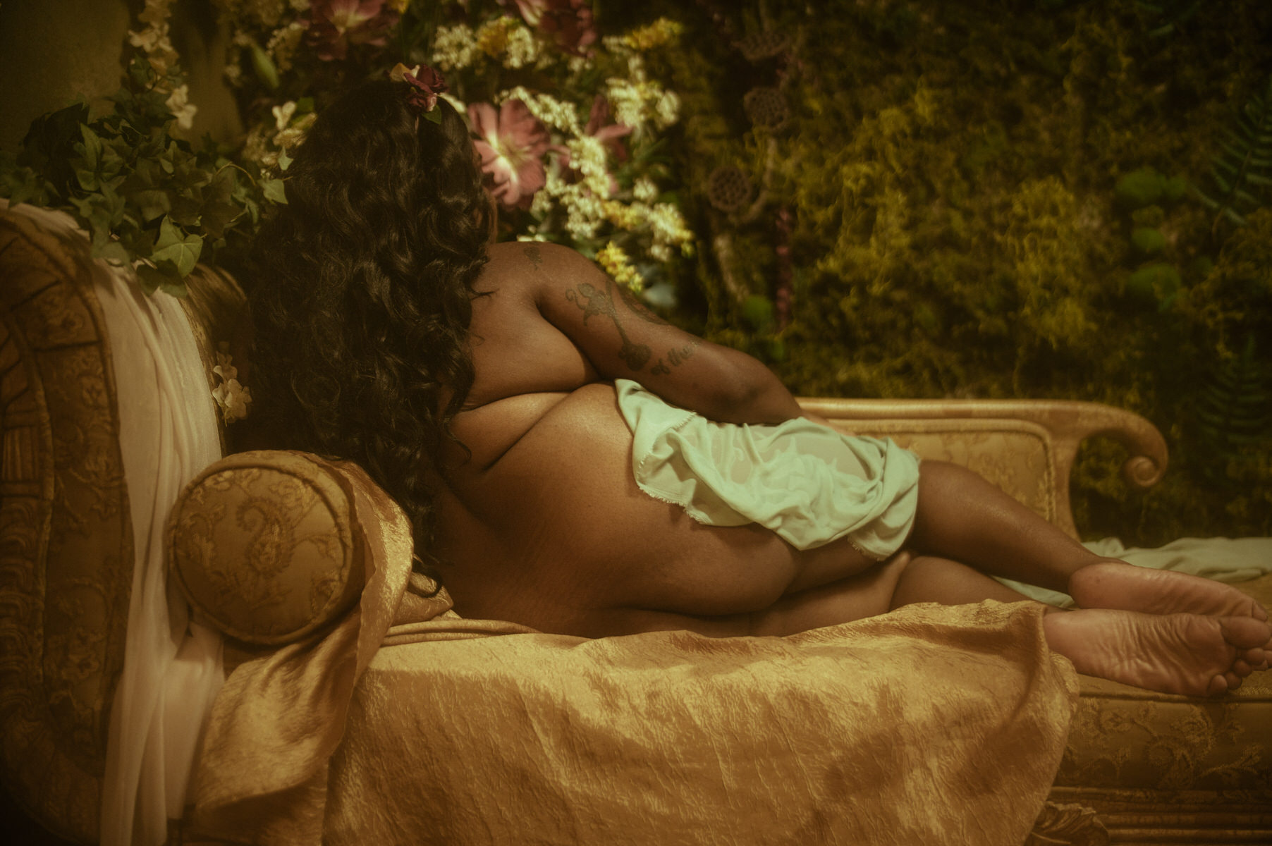 A nude woman sitting on a couch in front of flowers.