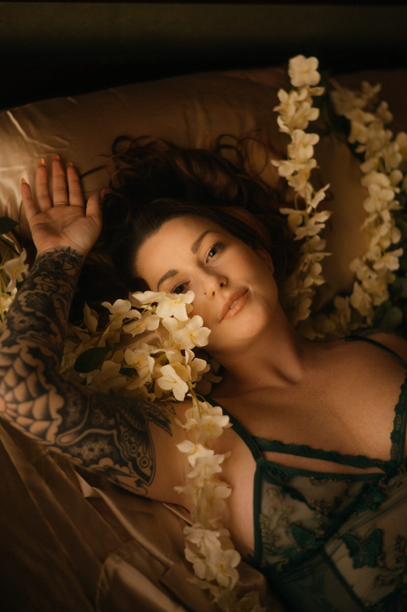 A woman with tattoos posing sensually in bed surrounded by beautiful flowers.