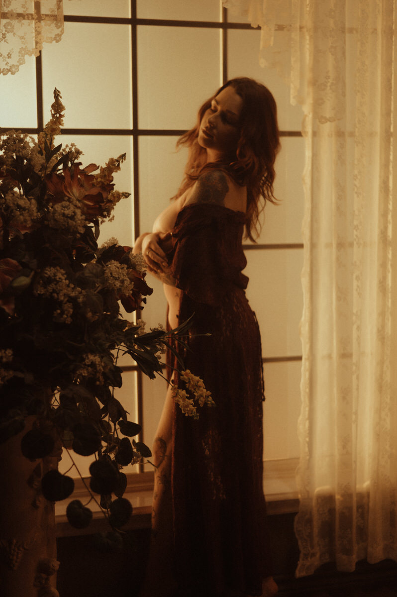 A woman in a dress posing in front of a window, showcasing elegant boudoir photography.