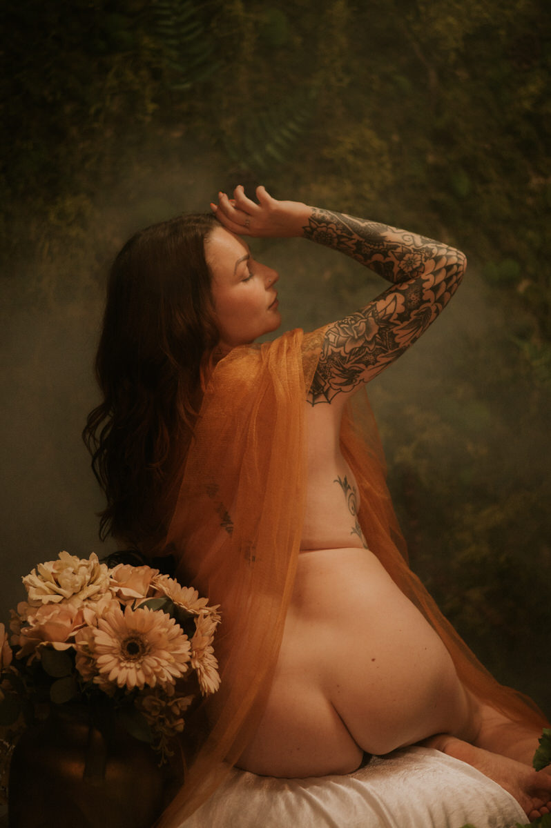 An artistic boudoir photograph capturing a nude woman with tattoos in a forest.