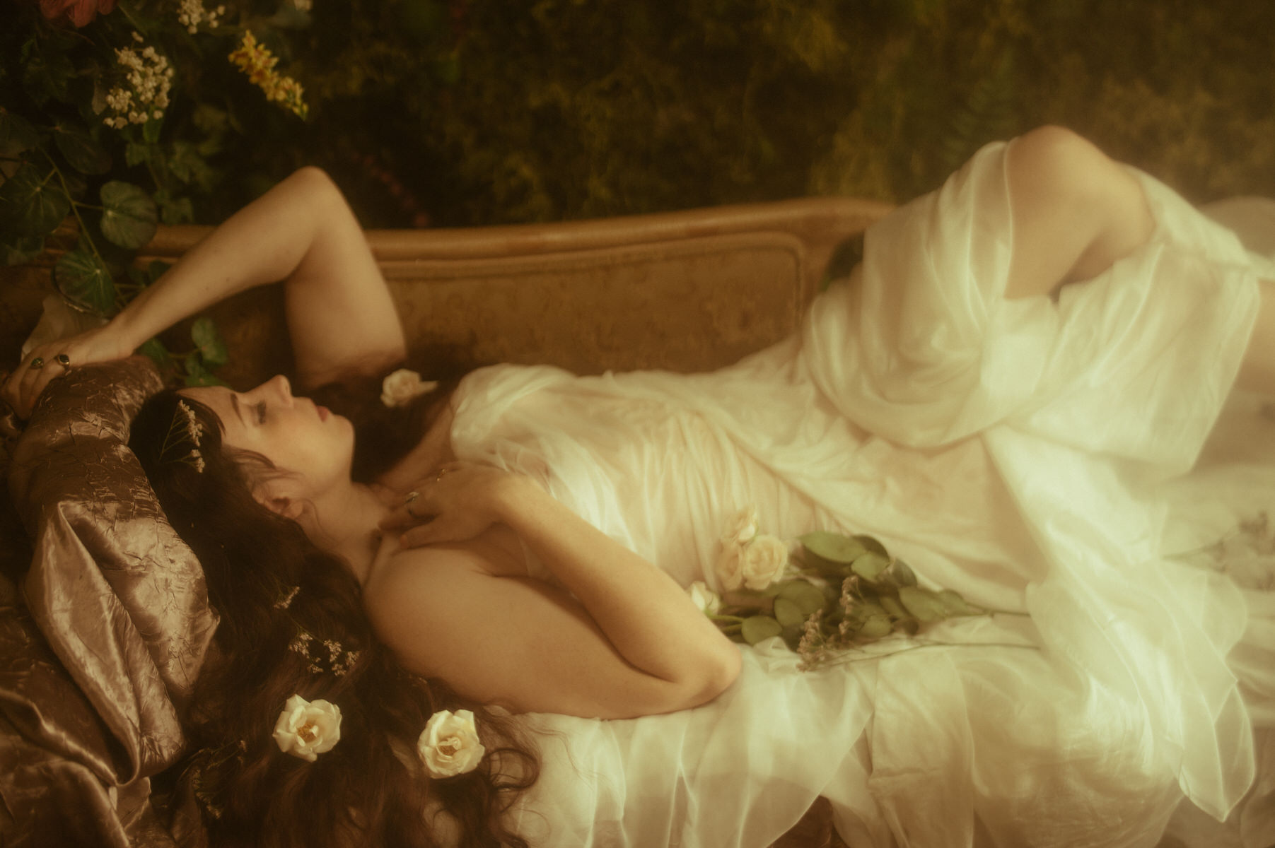 A dreamy woman in a white dress lounges on a couch in a boudoir setting.