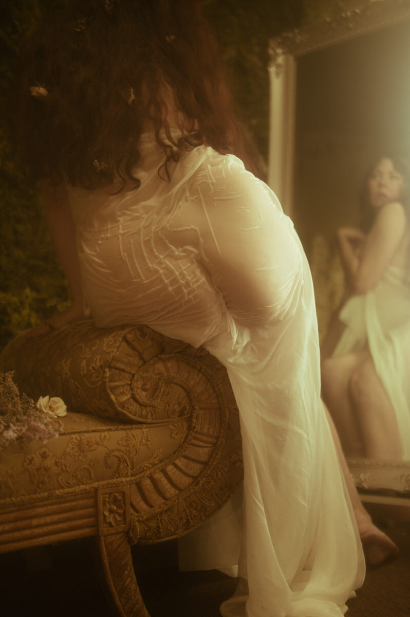 A woman in a white dress, with a wet fabric look, is posing in front of a mirror for her boudoir photography session.