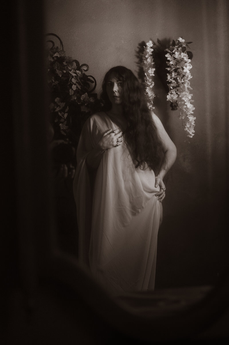 A woman in a dark and moody boudoir setting, wearing a white dress, stands elegantly in front of a mirror.