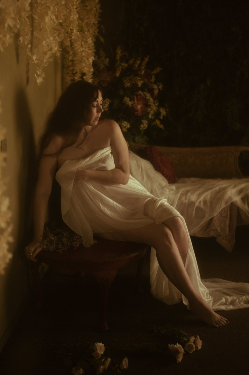 A women covered in white silky fabric posing on a chair for a moody boudoir photoshoot.