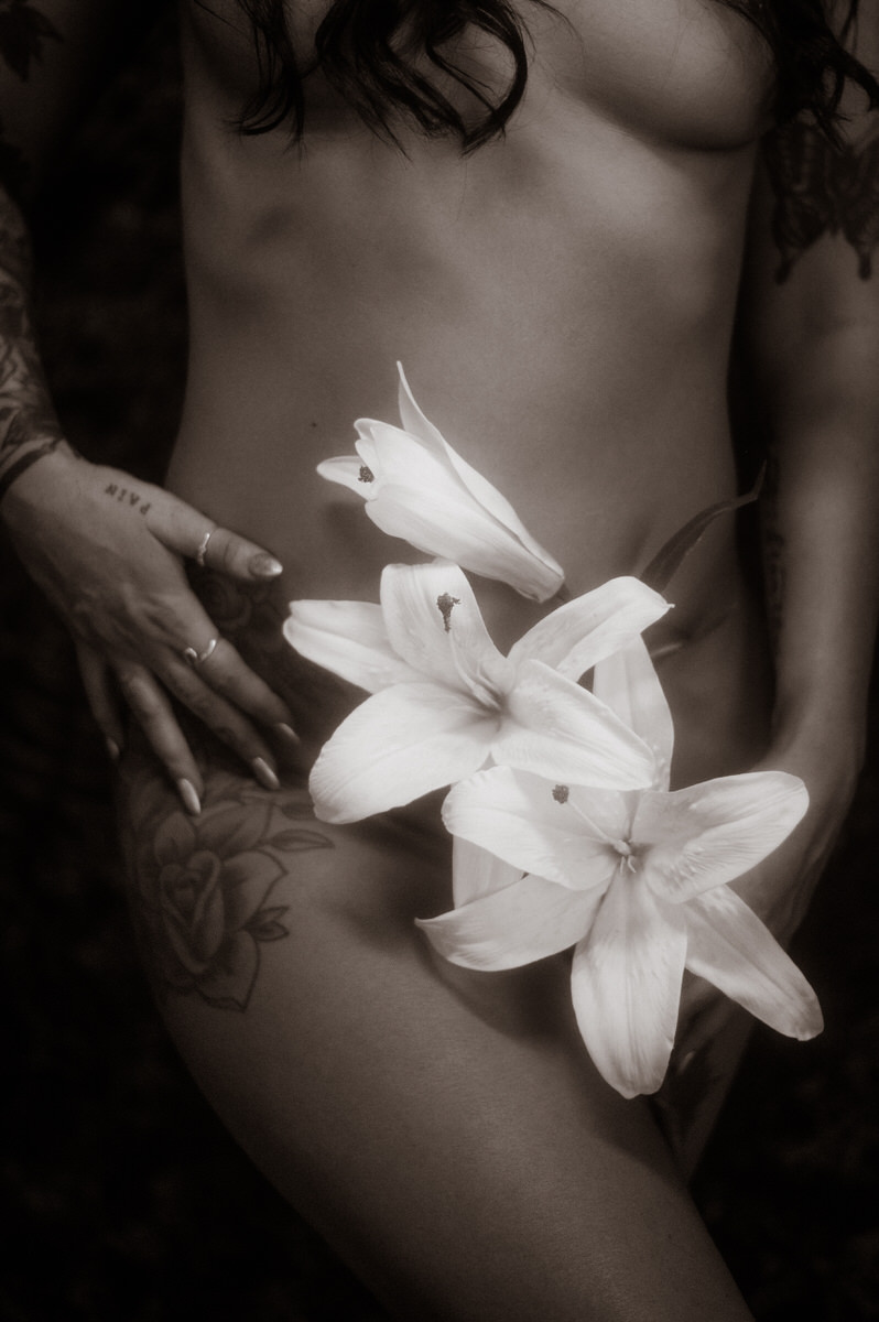 A nude woman with tattoos posing for boudoir photos, delicately holding flowers.