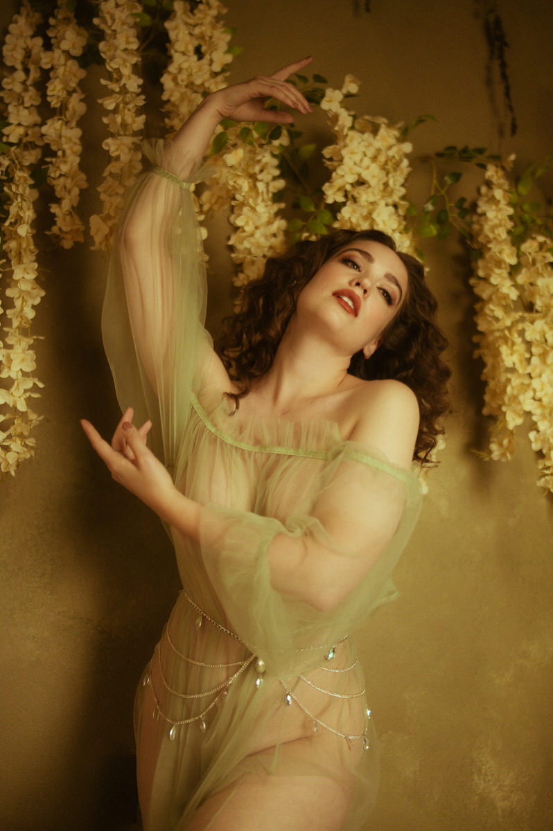 A woman in a green duster striking a painterly boudoir pose among flowers.
