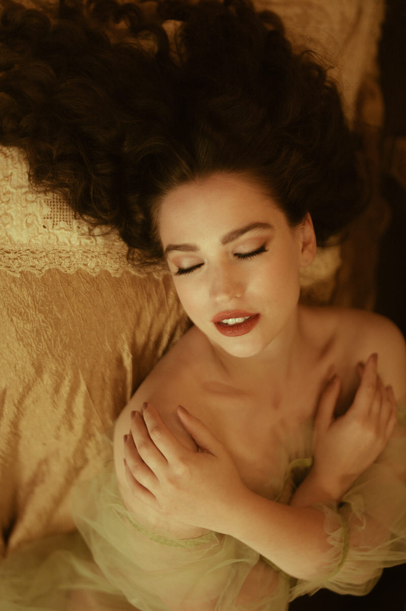A woman with long hair in a pre-Raphaelite-inspired pose, reclining on a bed in a fine art boudoir setting.