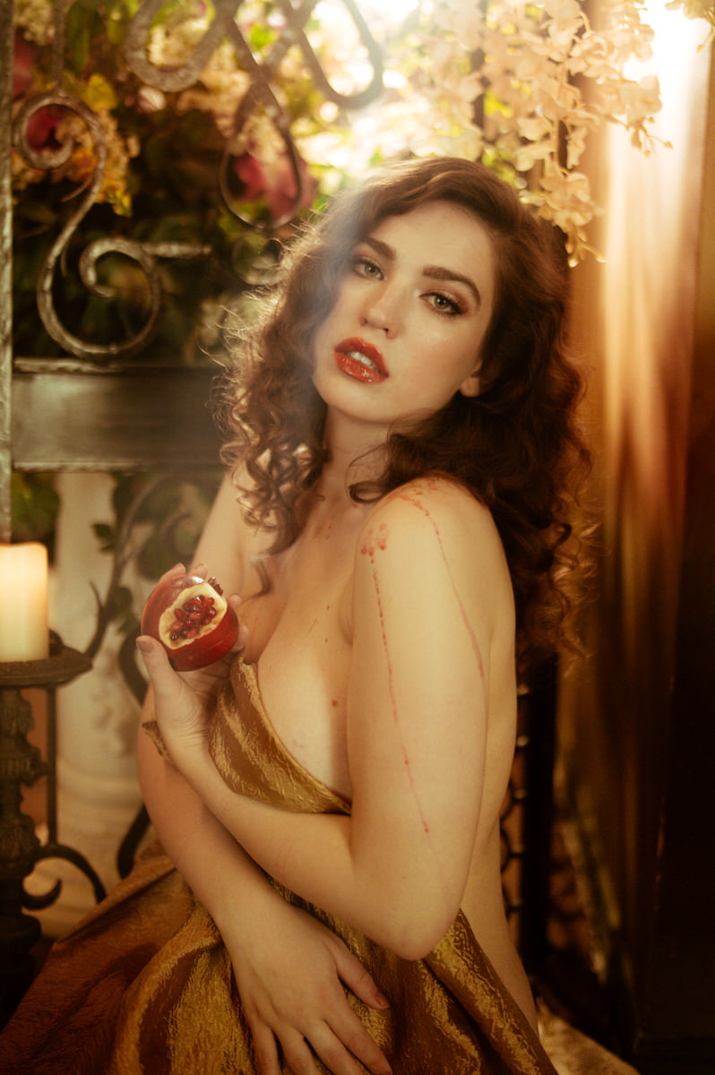 A woman in a golden dress elegantly holds a pomegranate, embodying the delicate allure of fine art boudoir.