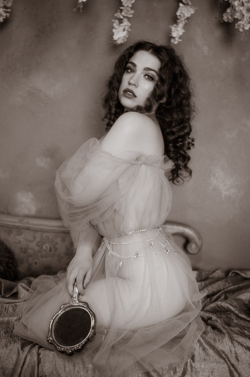 A woman in a sequined dress, exuding the essence of pre-Raphaelite beauty, captivatingly sits on a bed in this stunning fine art boudoir photograph taken in Dallas.