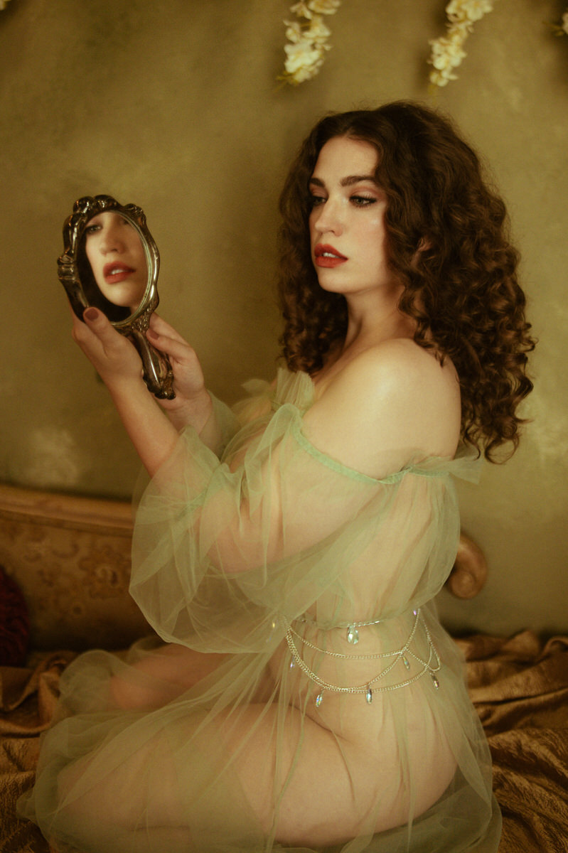 A woman is sitting on a bed looking at herself in a vintage hand mirror for a boudoir photoshoot inspired by pre-Raphaelite paintings.