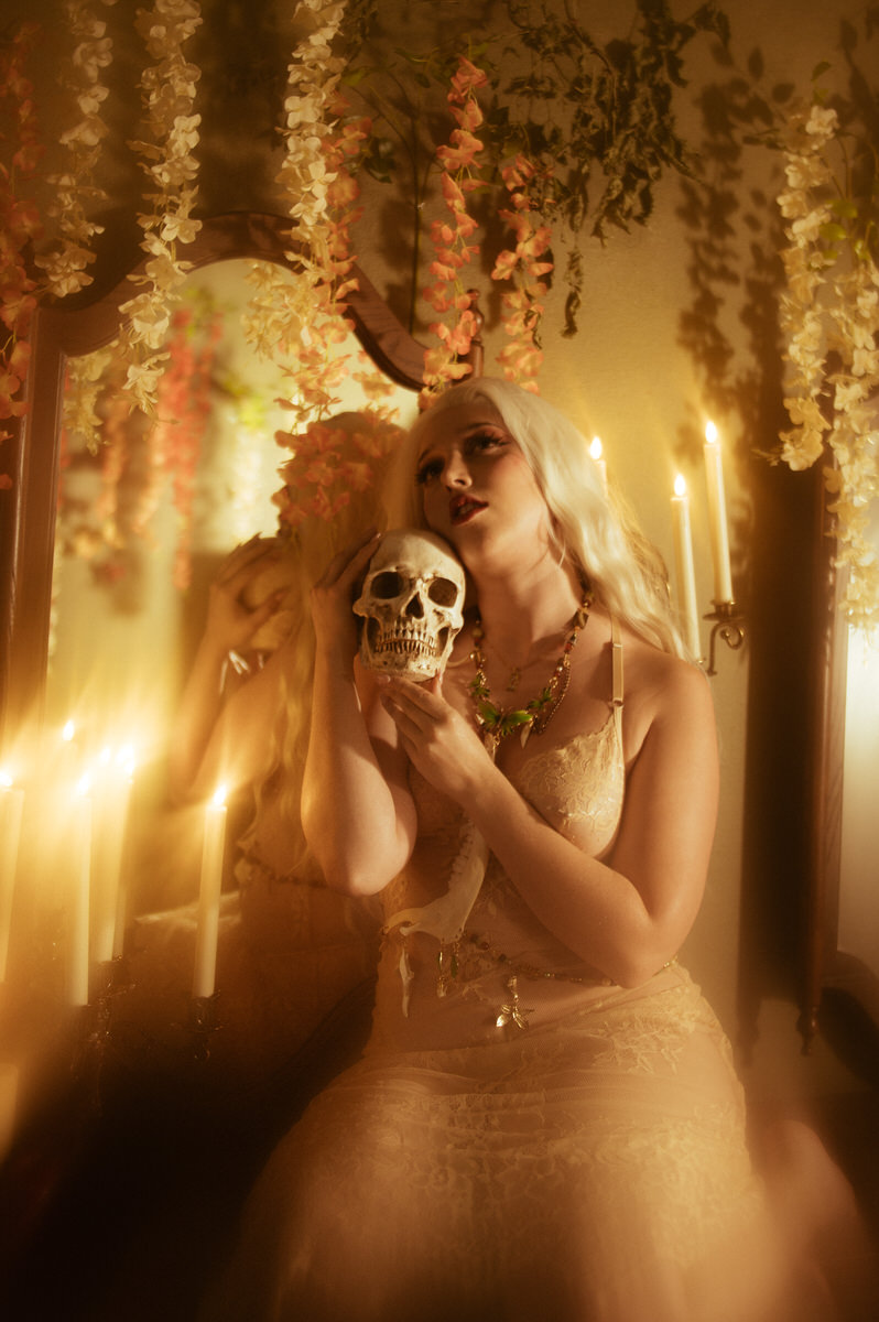 A witchy woman gazes at her reflection in a mirror while holding a skull.