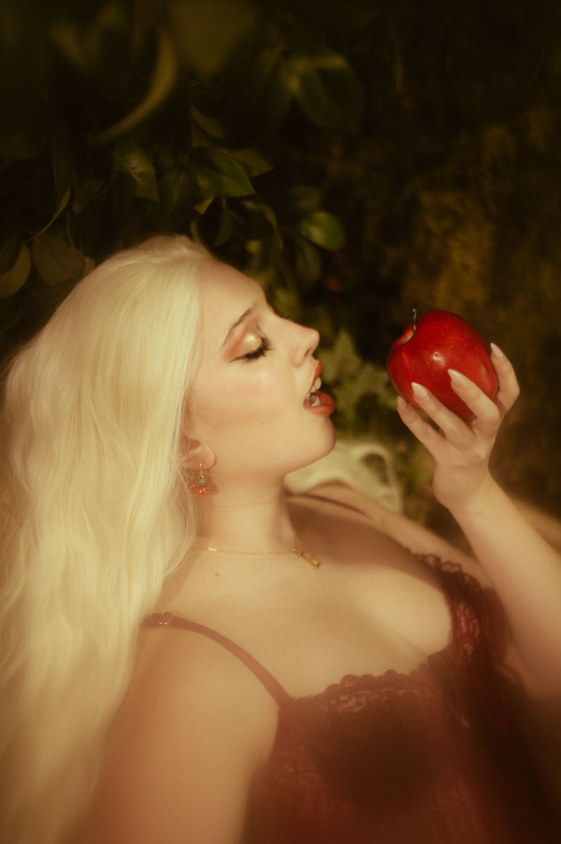 A woman in a red dress gazes sensually at the camera as she eats an apple.