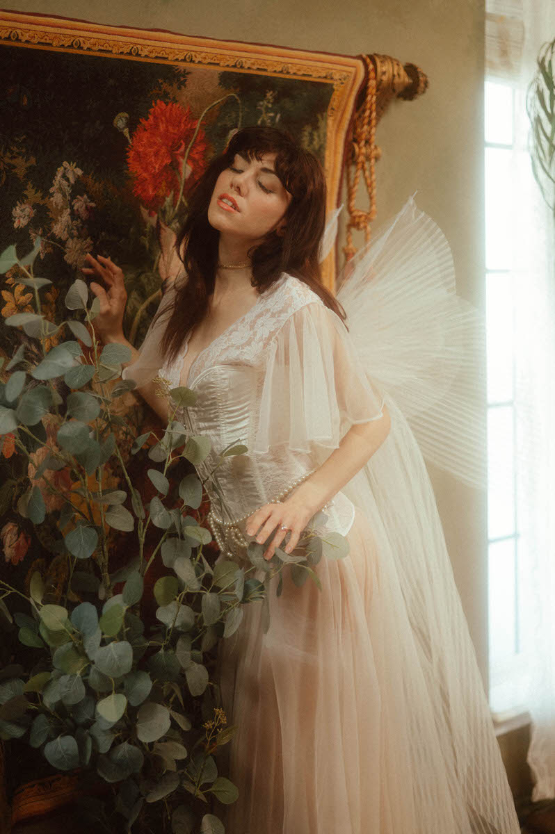 A person in a light and airy costume with angel wings poses beside a painting and foliage, evoking a fantasy ambiance.