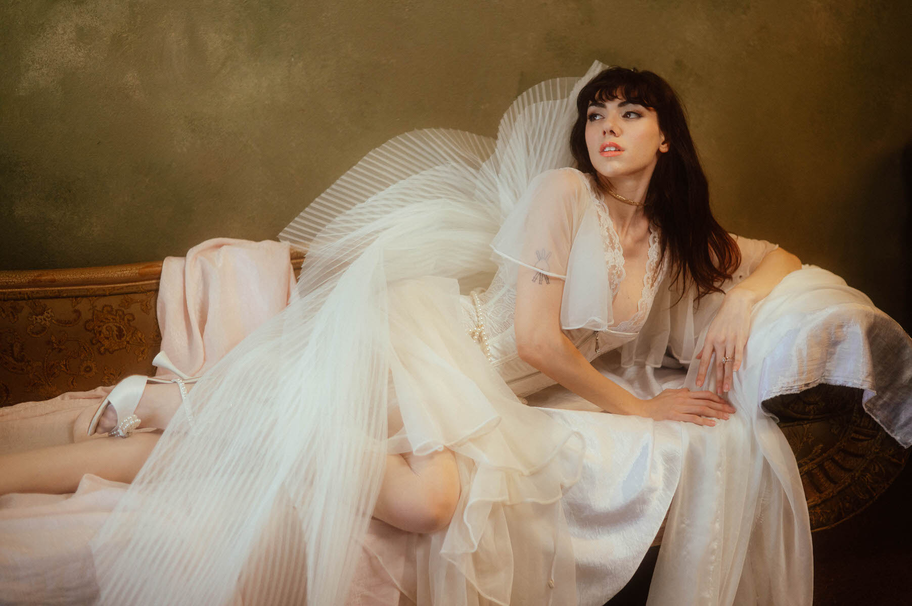 A woman in a vintage white robe outfit with angel wings reclining elegantly on an antique sofa against a green wall for a boudoir photoshoot.