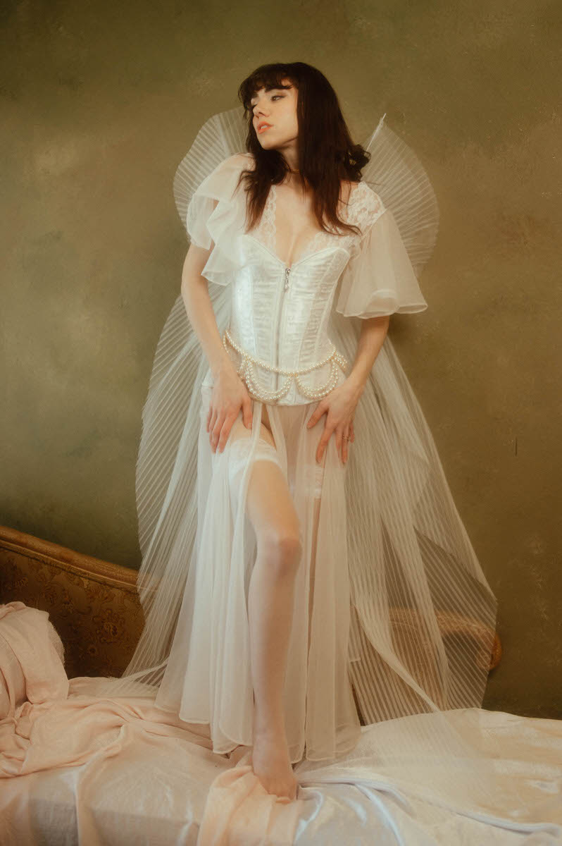 Woman in a vintage-inspired white corset and sheer robe, wearing a pair of tulle angel wings, posing against an olive green wall.