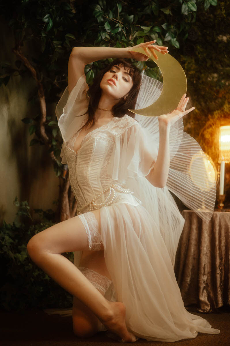 A woman in a flowing white dress and angel wings by Stonehard Jewelry, posing with a crescent moon prop in a dimly lit, plant-filled room.
