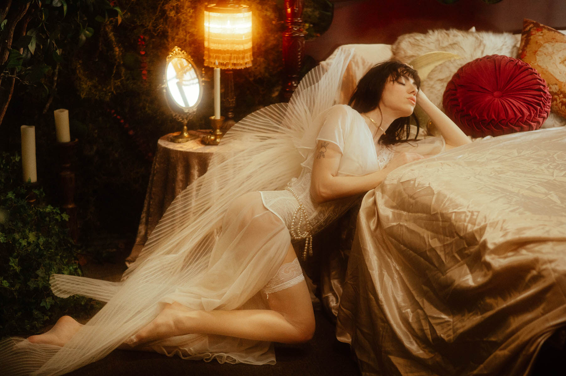 Woman in a sheer dress reclining on a bed in a Dallas boudoir studio with warm lighting and plants.
