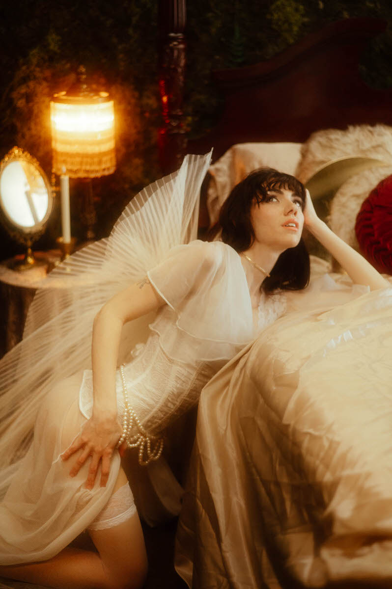 Woman in a vintage-style dress posing on a bed adorned with Stonehart Jewelry floating wings, creating a warm, soft-focus ambiance.