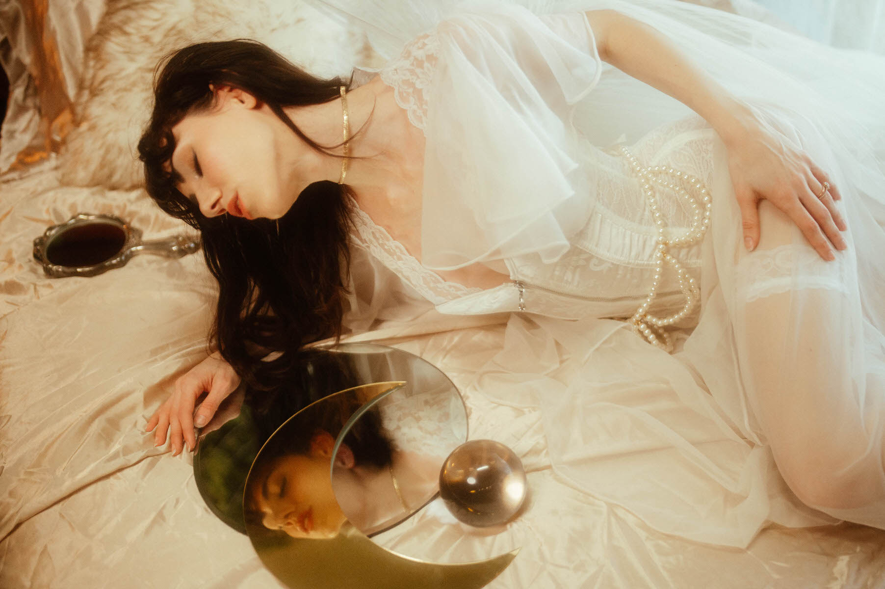 A woman dressed in a white gown and angel wings lies on a bed, her reflection visible in a curved mirror placed beside her.