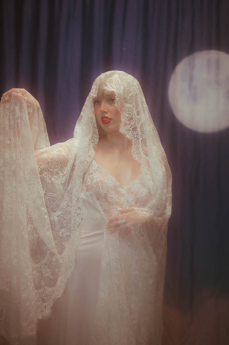 A woman in a lacy bridal gown and veil, posing elegantly for a winter boudoir shoot, with a soft focus and dreamy, full moon backdrop.