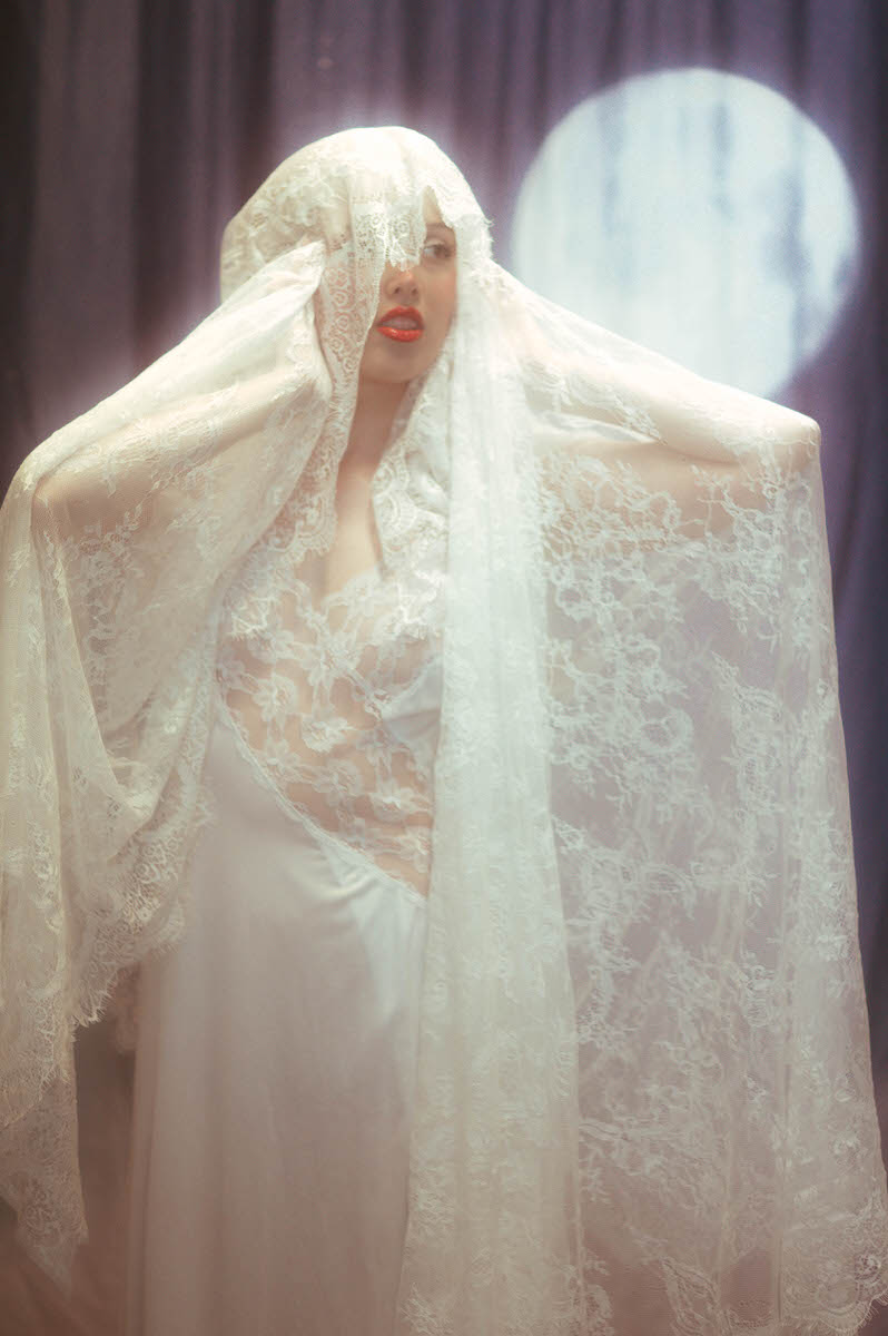 A woman in a lacy white veil and dress stands dramatically with a surprised expression, backlit by a glowing circular light during her Denton, TX boudoir photoshoot.