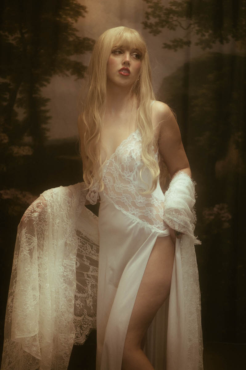 A woman in a white lacy dress and gloves poses behind a hazy glass pane, with a forest backdrop creating a dreamy, ethereal effect during her winter boudoir shoot.