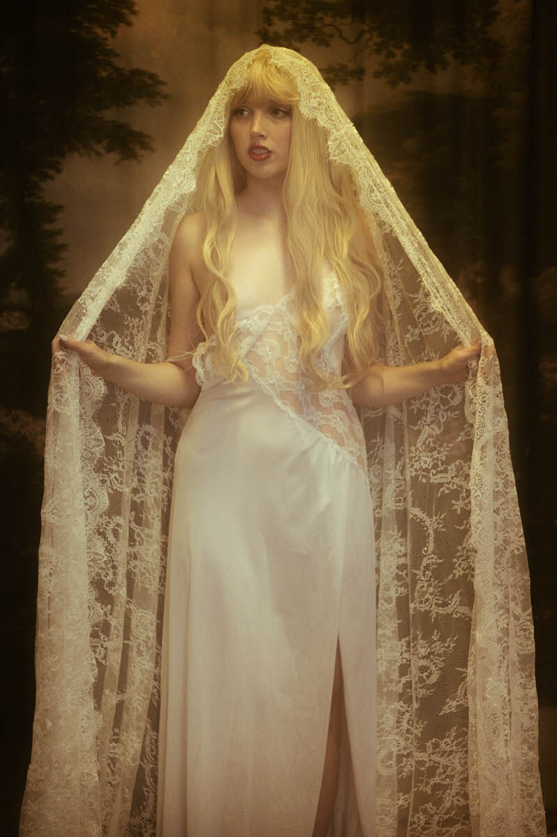 A woman with long blonde hair wearing a white lace gown and veil, standing in a misty forest setting during a winter boudoir shoot.