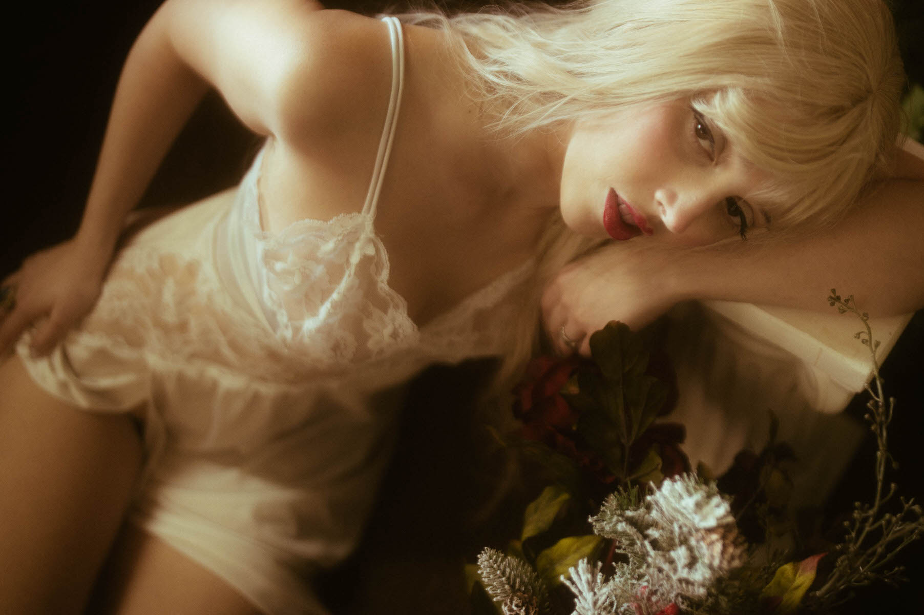 A woman in white lace lingerie reclining with flowers for a winter boudoir shoot, soft focus.