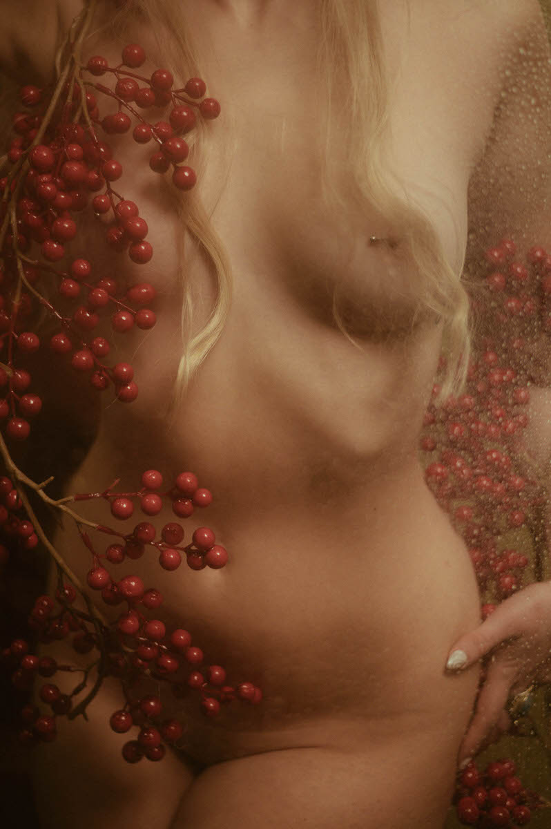 A nude woman posing in front of foggy glass, holding red berries, seen through a translucent, textured surface during a winter boudoir shoot.