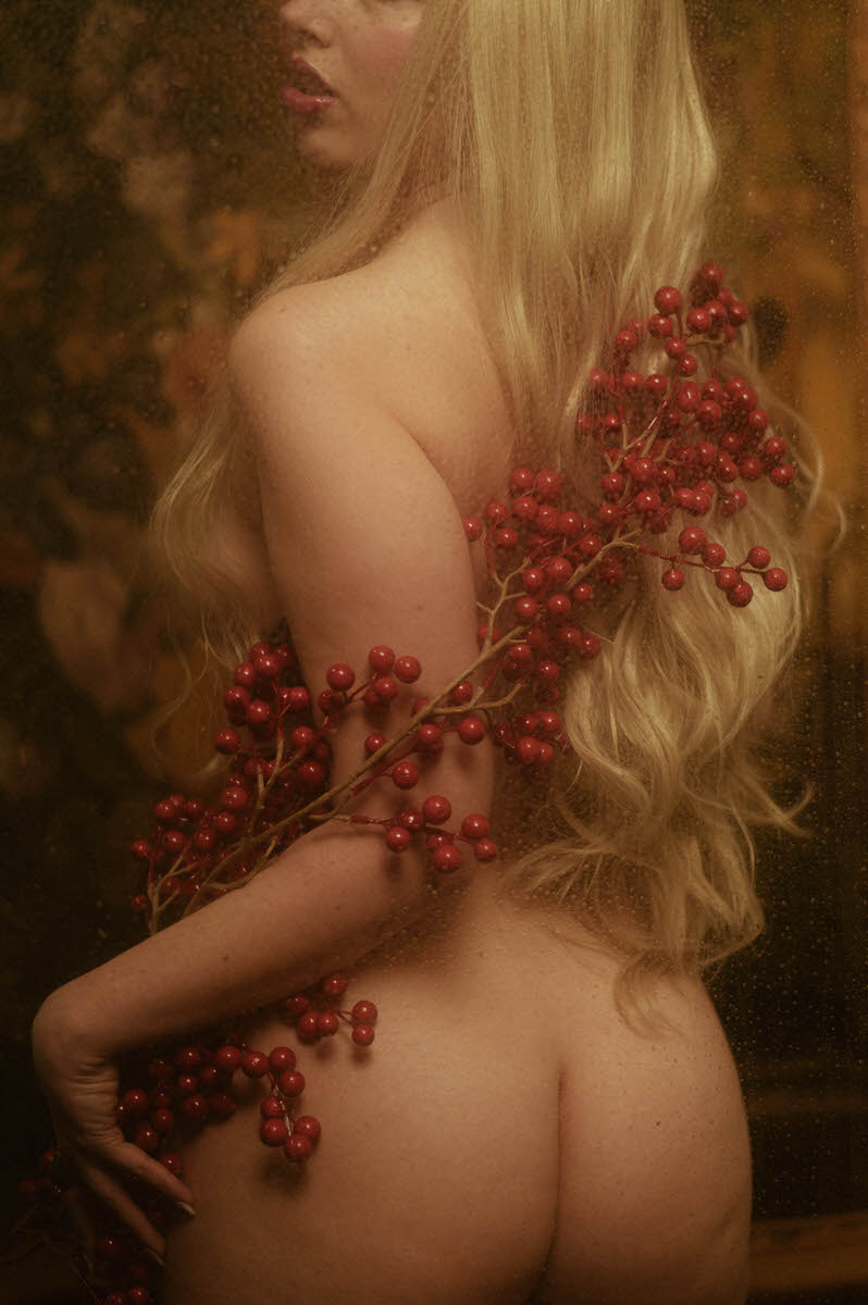 A woman with long blonde hair holds branches with red berries during a yule photoshoot, her back to the camera, in a softly focused, warm-toned setting.