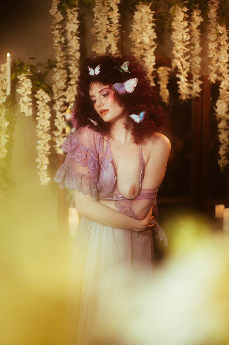 A woman with curly hair adorned with butterflies is wearing a sheer purple garment, standing in front of hanging floral decorations, creating a whimsical fairy tale photography scene in Dallas TX.