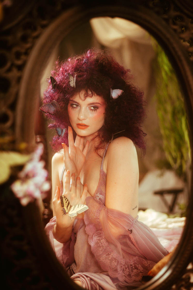 A woman with curly purple hair adorned with butterflies gazes into a mirror, holding a butterfly in their hands, dressed in a lavender lace outfit, creating an enchanting scene reminiscent of fairy tale photography.