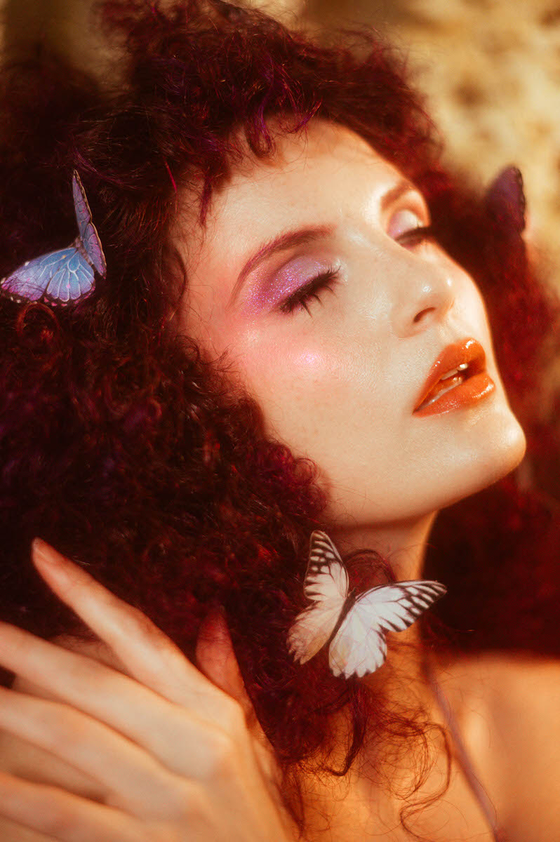 A woman with curly hair, adorned with purple eyeshadow and orange lipstick, stands gracefully in a whimsical butterfly photoshoot, where two delicate butterflies rest on their hair and hand.