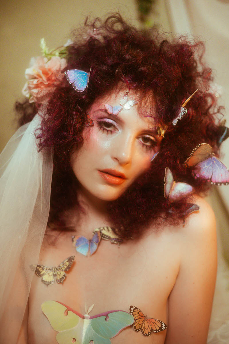 A woman with curly hair adorned with colorful butterflies and flowers, wearing makeup and a sheer veil, poses looking into the camera, embracing the essence of a spring boudoir session in Dallas.