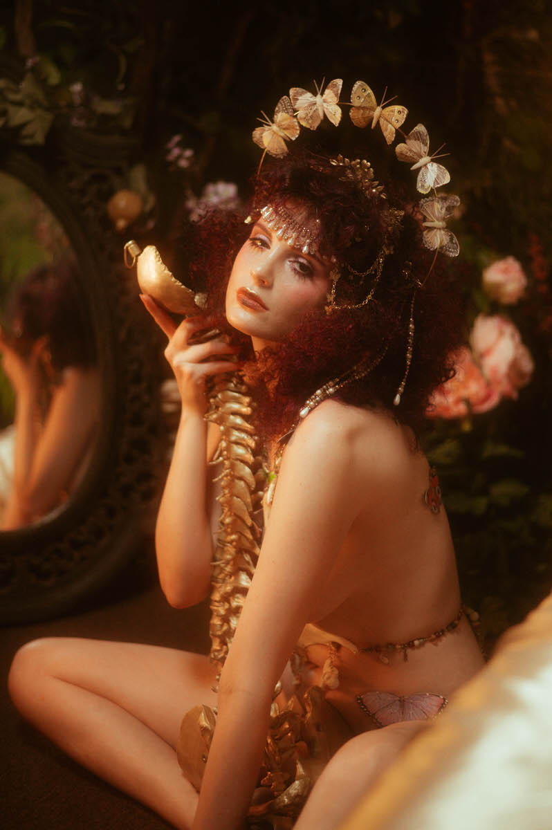 A woman with curly hair adorned with flowers and butterflies, holding a gold faux spine, sitting in front of a mirror surrounded by flowers—a perfect scene for a spring boudoir butterfly photoshoot in Dallas, TX.