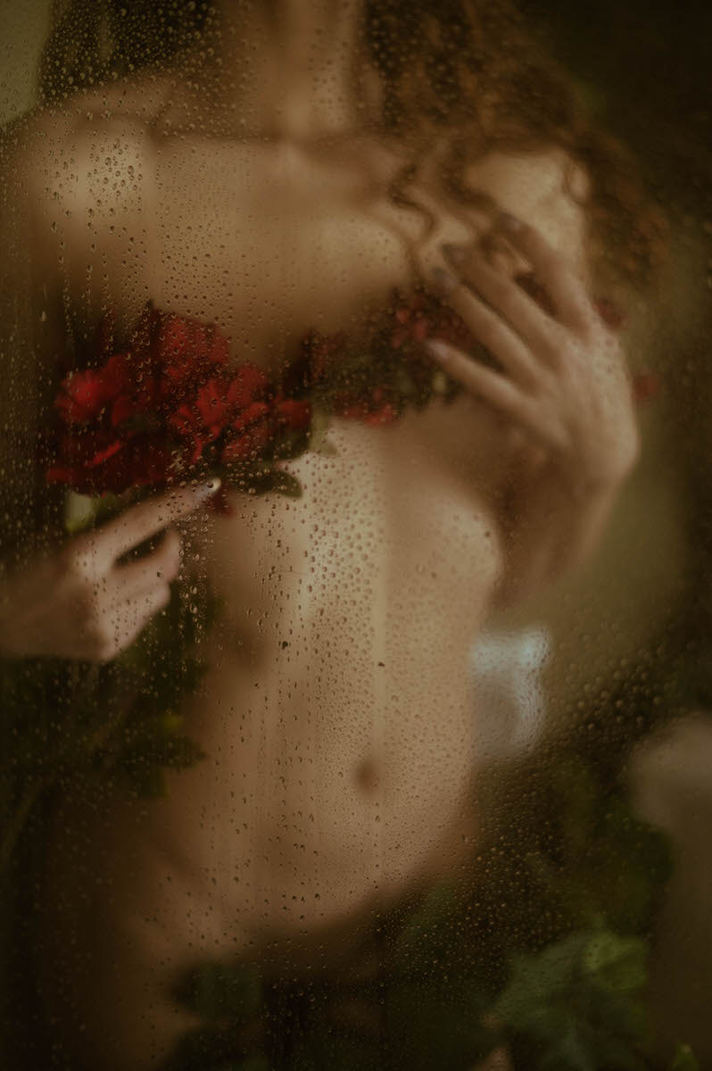 A fine art nude photography piece captures a partially obscured figure with long hair, holding red roses against their chest behind a glass surface covered in water droplets, evoking the essence of a faux shower boudoir.