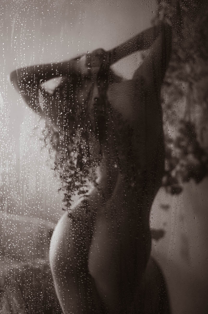 A silhouette of a person standing behind a fogged glass door with water droplets, striking an arched pose with hands on the head in a dimly lit environment evokes the allure of a faux shower boudoir shoot.