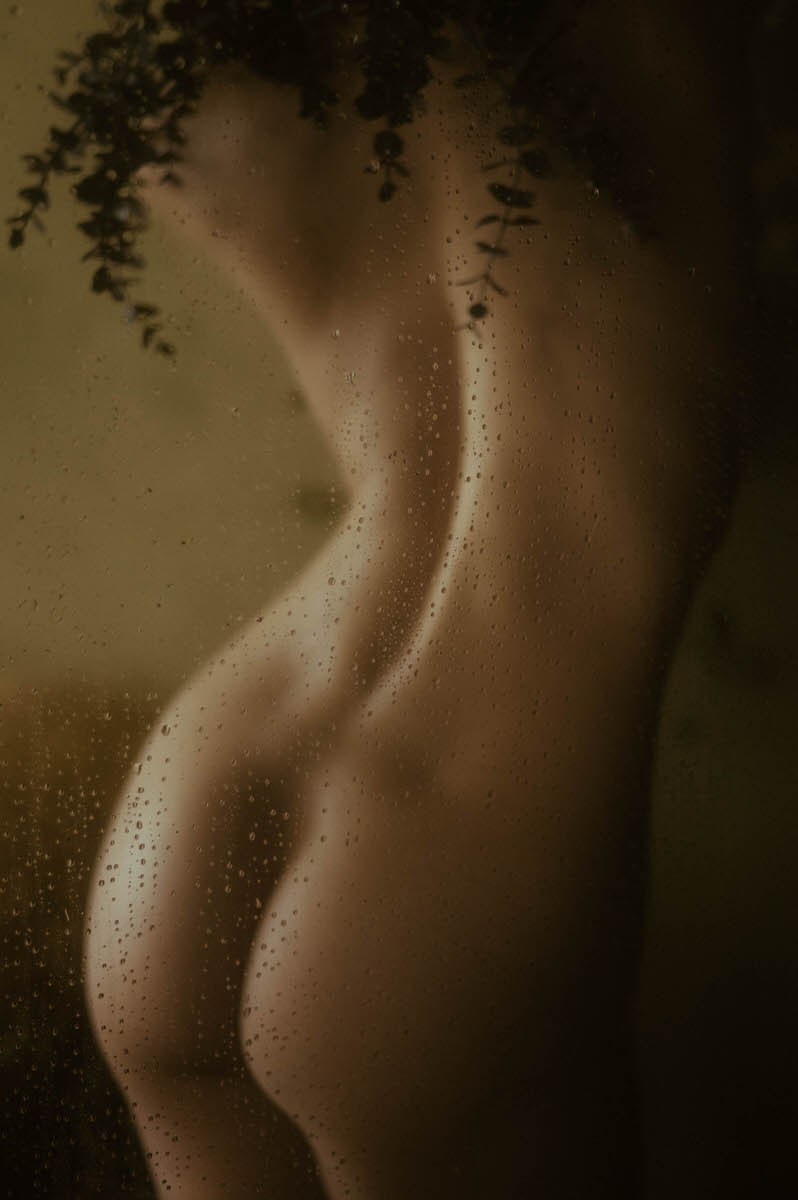 A nude figure is partially visible behind a fogged glass pane with water droplets. This fine art boudoir setting highlights the silhouette, emphasizing the curves of the body in a dimly lit atmosphere.