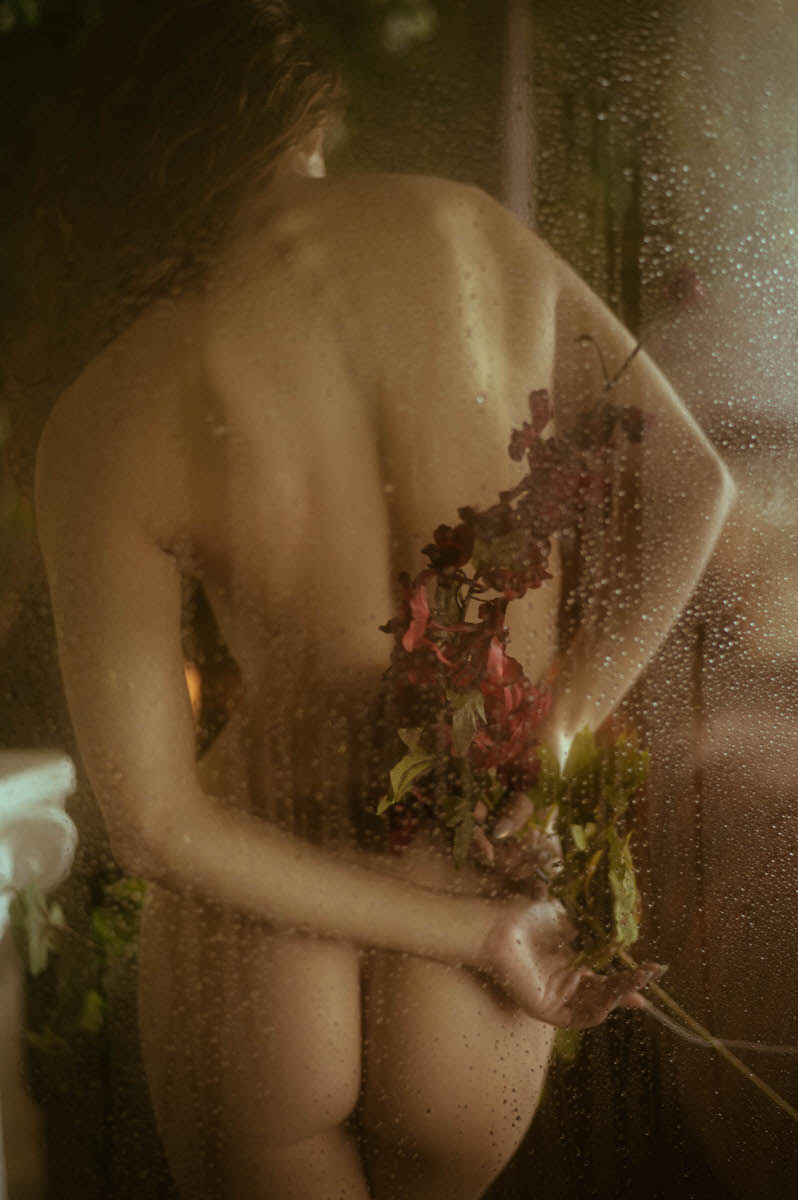 A person, seen from behind, stands nude holding a bouquet of wilted flowers in one hand. The scene, like a fine art boudoir photograph, is blurred through a wet glass surface with visible water droplets.