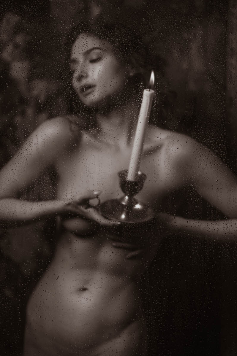 A nude woman holds a lit candle while standing behind a wet glass surface, evoking the intimate atmosphere often seen in fine art nude photography, in a dark, moody, black-and-white photograph.