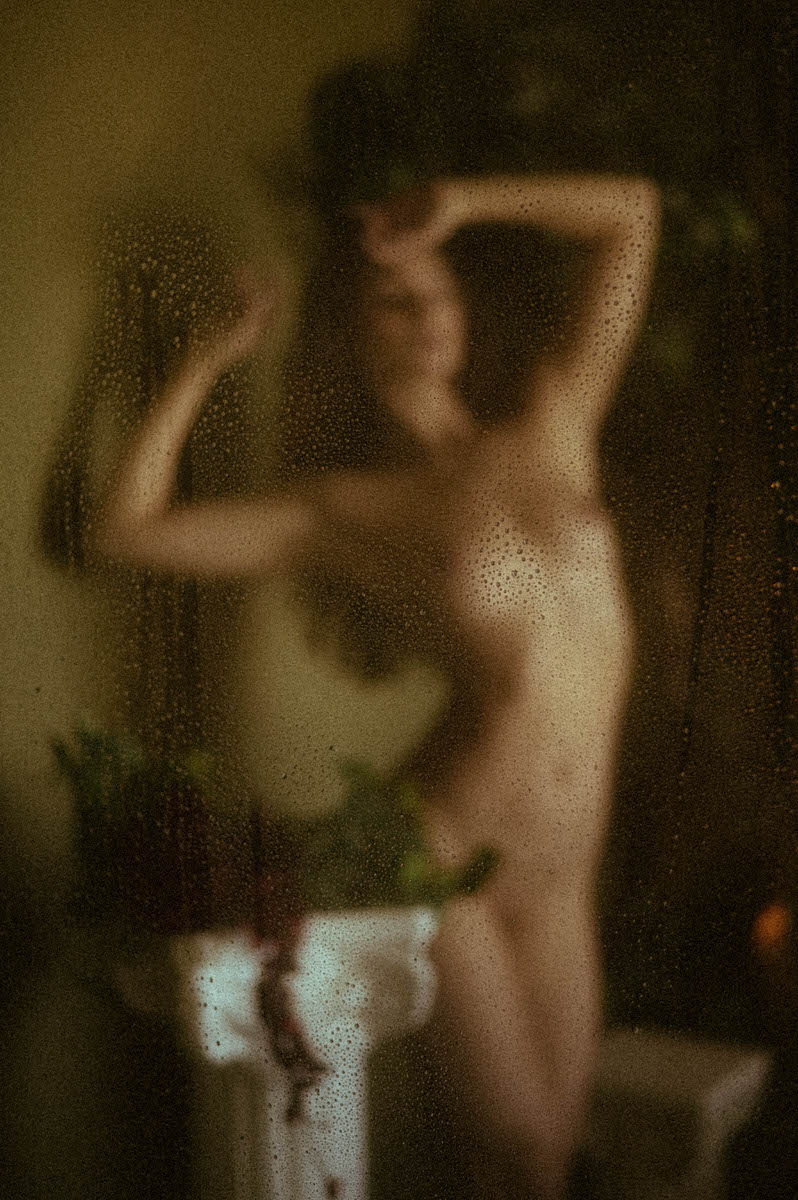 Blurry image of a nude person standing behind a foggy, water-speckled glass. They have one hand raised to their head and the other bent at the elbow, creating a soft, obscured silhouette reminiscent of fine art boudoir photography.