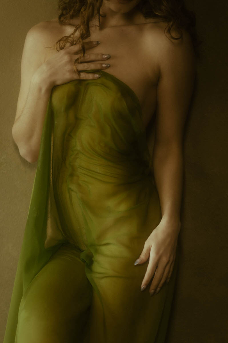 A woman partially covered with a green, wet sheer fabric stands against a tan background with one hand on their neck and the other by their side, creating an evocative faux shower boudoir vibe.