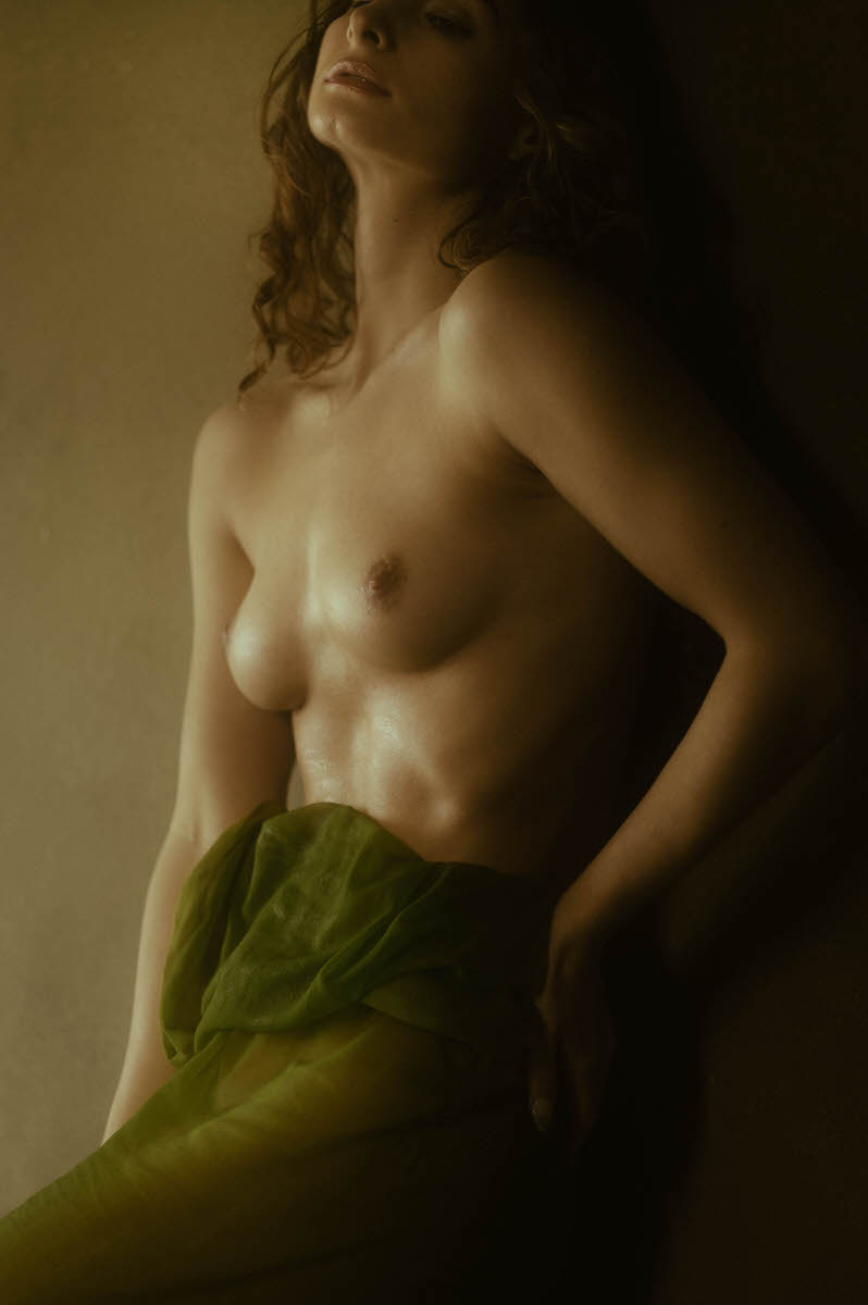 A topless woman with light skin stands against a wall, partially wrapped in a green wet fabric around her waist. She has curly hair and a contemplative expression in this fine art boudoir image, which is enhanced by soft, warm lighting.