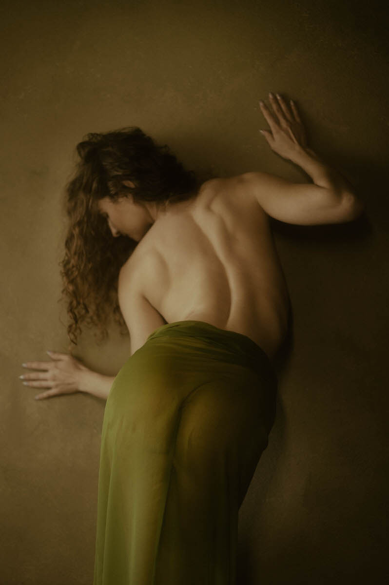 A woman with long, wavy hair stands against a wall, partially draped in a green, semi-transparent fabric that covers their lower body. Their upper body is bare, and their back is facing the camera in an exquisite fine art nude photography moment.