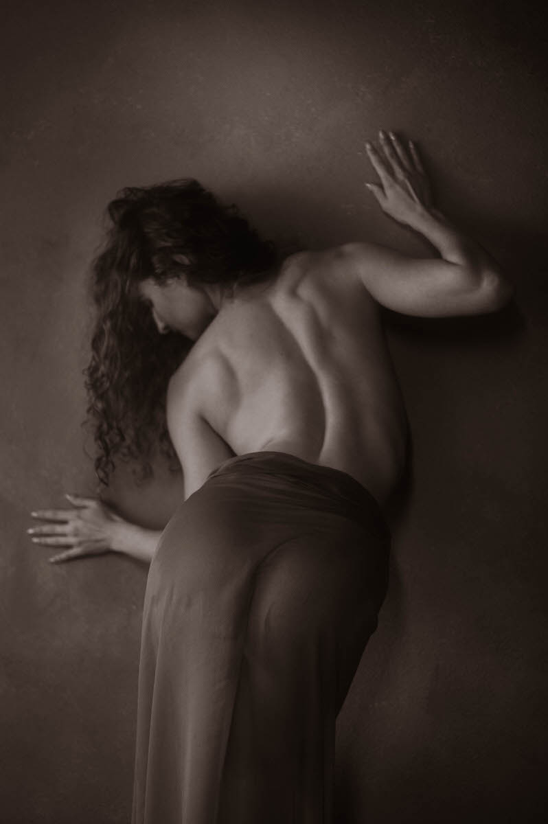 A monochrome image of a woman with long curly hair pressing against a wall, viewed from the back. The figure is partially draped in a sheer, wet fabric; it captures the essence of fine art boudoir photography.